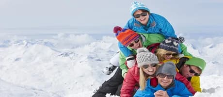 A cheerful group of skiers in colorful jackets and beanies pile together and smile at the top of a snowy summit, celebrating a day on the mountain as a group.