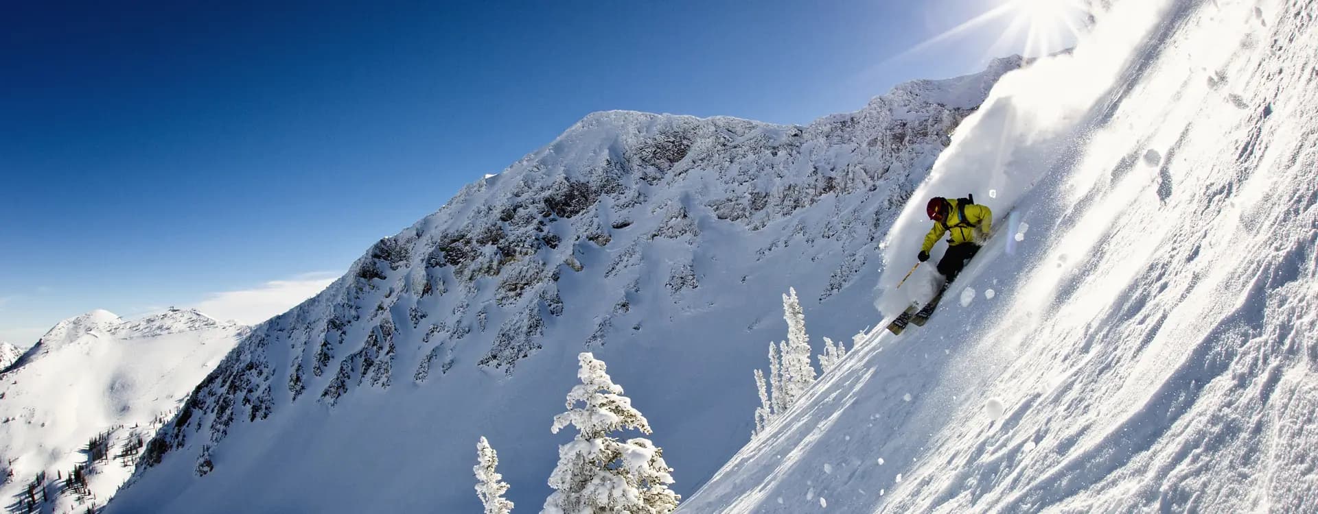 A skier in a bright yellow jacket carves down a steep powder slope beneath a jagged, snow-covered peak, kicking up a plume of snow with the sun flaring overhead against a deep blue sky.