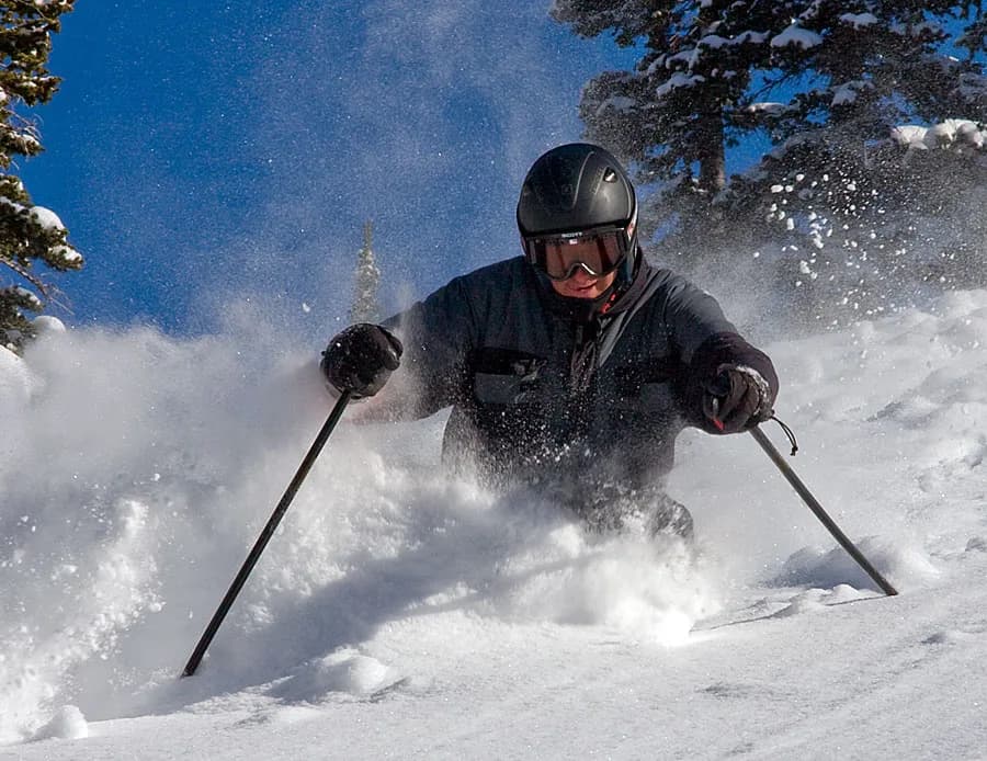 A skier in a black helmet, goggles, and dark jacket skis directly toward the camera through deep powder, snow spraying up around their knees on a bluebird day.