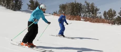 An instructor in a blue jacket demonstrates a turn on a gentle groomed run while a student in blue follows close behind, in a one-on-one private lesson.